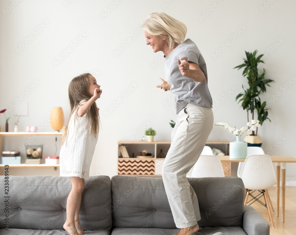 Cute little girl having fun playing with smiling grandmother jumping on couch together, happy granny and active kid grandchild dancing on sofa, grandma and granddaughter laughing playing at home