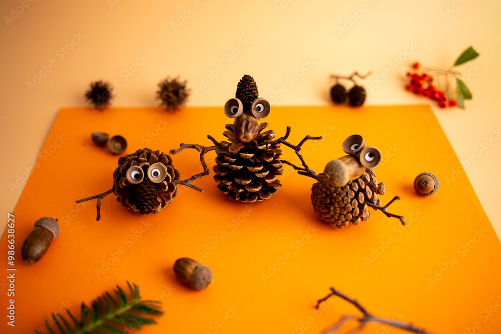 Creative arrangement of pine cones with googly eyes, twig limbs, and acorns on an orange background, resembling playful creatures in a whimsical nature scene,