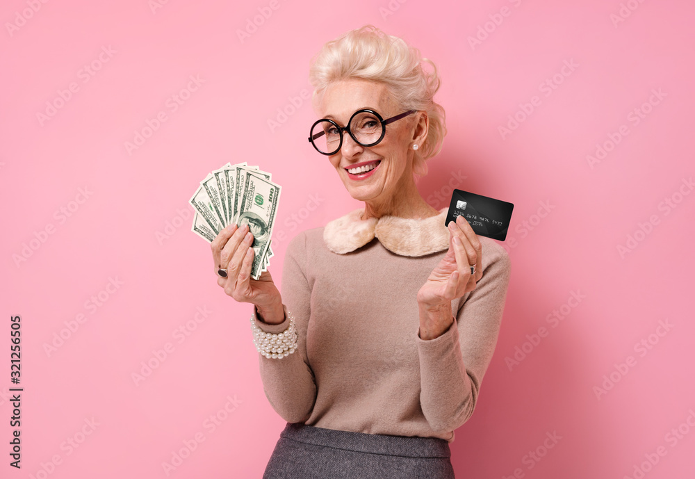 Smiling grandmother shows money cash and credit card. Photo of kind elderly woman on pink background.